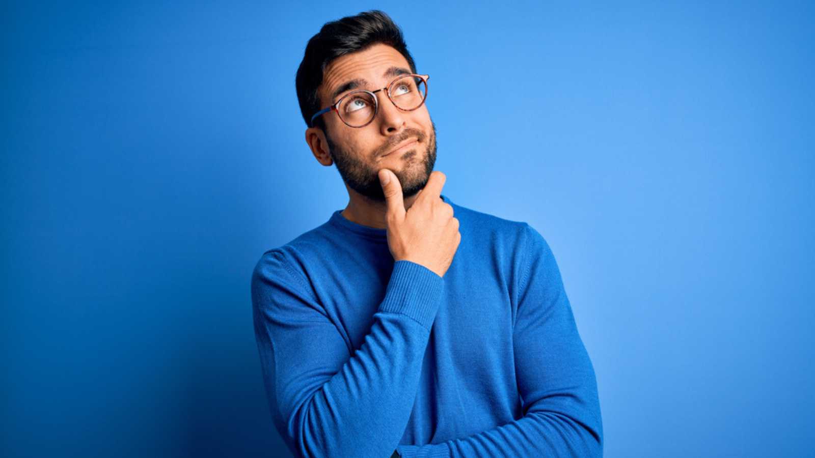Người đàn ông trẻ có râu đẹp trai mặc áo len giản dị và đeo kính trên nền xanh đang suy nghĩ về việc lo lắng về một vấn đề nào đó và đặt tay lên cằm cảm thấy lo lắng và hồi hộp